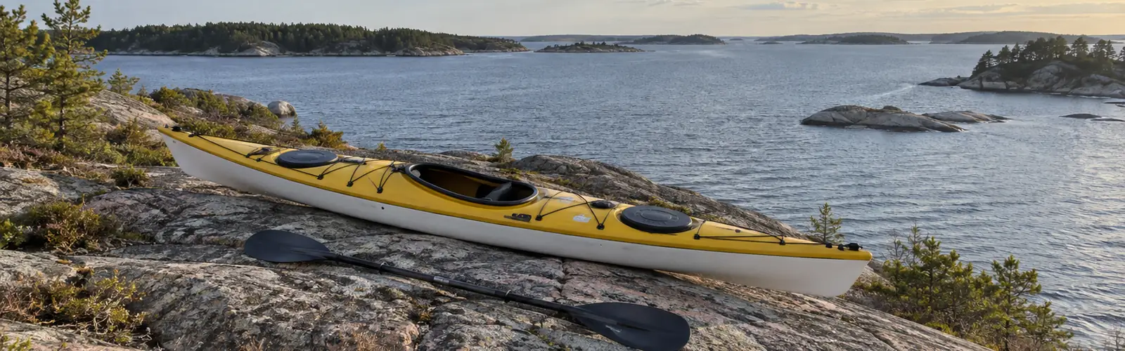 Gul och vit havskajak med paddel på en granithäll, med en lång rad skärgårdsöar i fjärran