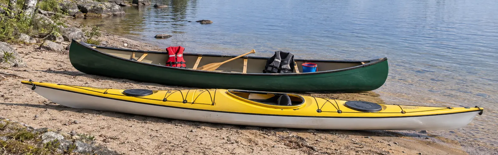 Grön kanot med en liten röd flytväst, en kort paddel och en hink, bredvid en gul och vit havskajak på en sandstrand vid en sjö