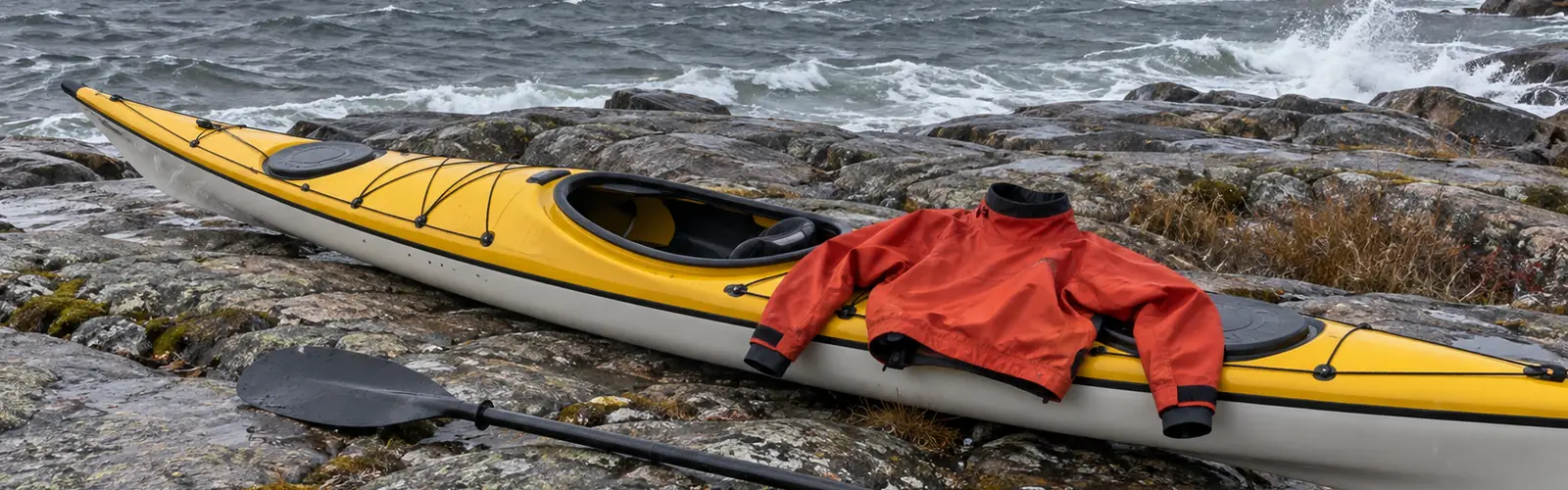 Röd paddeljacka med tättslutande hals och handledsmuddar utlagd på en havskajak på klipporna en blåsig höstdag