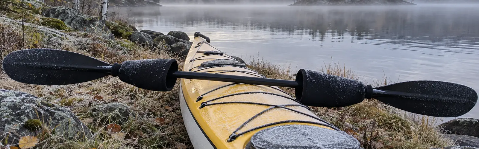 Paddel med pogies i neopren på skaftet, lagd över en frostig havskajak vid en dimmig sjö en höstmorgon