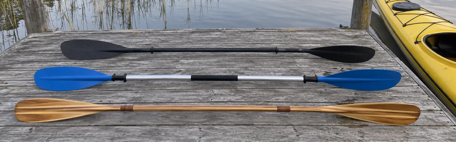 Tre kajakpaddlar i kolfiber, aluminium med blå blad och trä, utlagda på en brygga bredvid en gul kajak