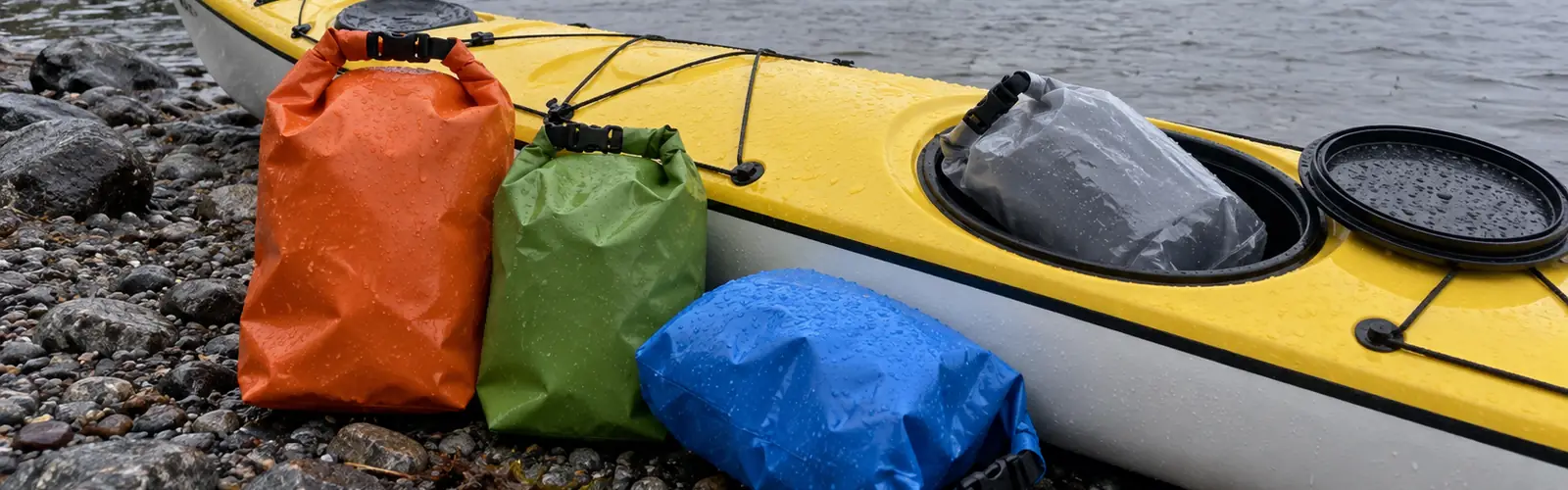 Orange, grön, blå och genomskinlig packpåse med regndroppar vid en havskajak med öppen lucka på en stenstrand