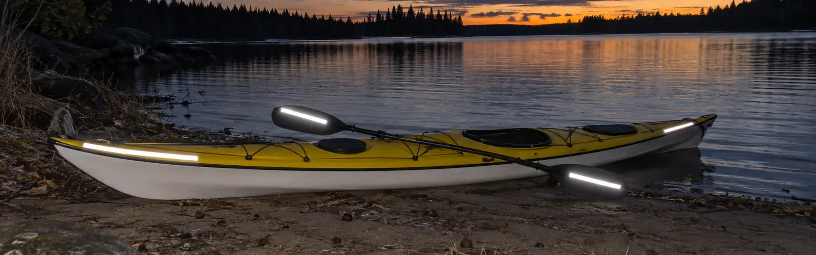 Havskajak och paddel med reflextejp som lyser i blixtljuset på en sjöstrand i skymningen